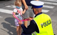 Przy przejściu dla pieszych, policjant daje opaskę odblaskowa małej dziewczynce, dziewczynka trzyma kobietę za rękę