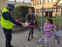 Policjant ruchu drogowego daje opaski odblaskowe dziewczynce na rowerze i kobiecie, dzieje się to obok szkoły
