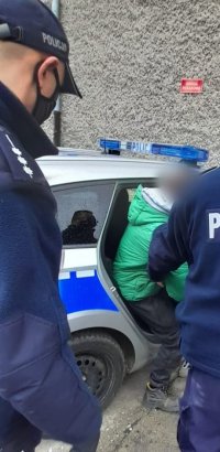 dwaj policjanci,zatrzymany mężczyzna w zielonej kurtce i ciemnych spodniach, radiowóz
