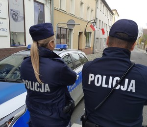 policjantka i policjant w tle budynki, na których wywieszone są flagi państwowe i radiwóz