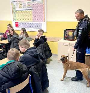 uczniowie siedzący w szkolnych ławkach, za nimi stoi policjant z psem służbowym