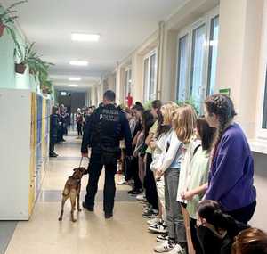 uczniowie na korytarzu, w tle szafki szkolne, okna, policjant z psem służbowym przemieszcza się obok szafek