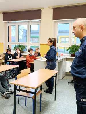 policjant i policjantka stoją zwróceni w kierunku uczniów, uczniowie siedzą w ławkach