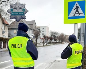 dwoje policjantów stojących obok jezdni, widoczne przejście dla pieszych i znak przejścia dla pieszych, w lewym górnym rogu gwiazda, logo krapkowickiej policji