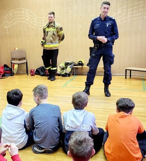 policjant, strażak i dzieci na sali gimnastycznej szkoły, policjant prowadzi prelekcję
