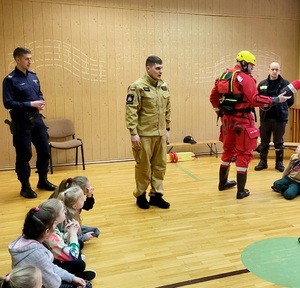 policjant i dwóch strażaków, prelekcja dla dzieci , dzieci siedzą tyłem do obiektywu na podłodze
