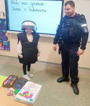 policjant i chłopiec ubrany w wyposażenia policyjne służące do ochrony ciała na zabezpieczeniach imprez masowych, hełm i kamizelkę