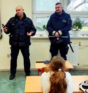 dwóch policjantów podczas prelekcji w klasie, na zdjęciu widoczna głowa rudowłosej dziewczynki siedzącej w ławce tyłem do obiektywu