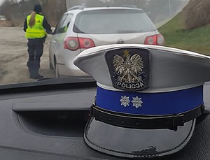 czapka policjanta ruchu drogowego na podszybiu samochodu, w tle zatrzymany do kontroli pojazd i policjantka kontrolująca kierowcę tego pojazdu