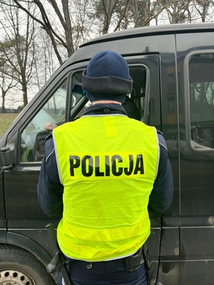 policjant kontrolujący pojazd koloru czarnego, stojący tyłem do obiektywu w umundurowaniu i żółtej kamizelce odblaskowej