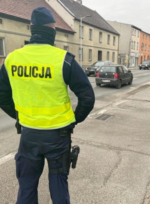 policjant nadzorujący ruch na terenie miasta, w tle budynki , jezdnia, dwa samochody osobowe, policjant umundurowany w żółtą kamizelkę odblaskową