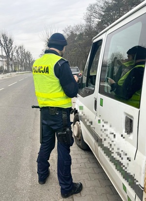 policjant kontrolujący pojazd koloru białego, stojący tyłem do obiektywu w umundurowaniu i żółtej kamizelce odblaskowej