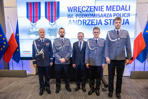 czterech policjantów w umundurowaniu galowym oraz mężczyzna w garniturze, w tle 2 flagi Polski i trzy flagi z symbolami Unii Europejskiej, dodatkowo widoczny ekran, na którym wyświetlone są obrazy medali