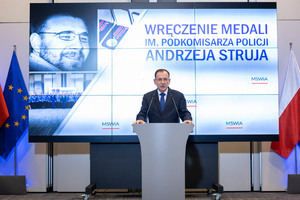 mężczyzna przy mównicy,Minister Spraw Wewnętrznych i Administracji Mariusz Kamiński, w tle ekran i flagi Polski oraz z symbolami Unii Europejskiej