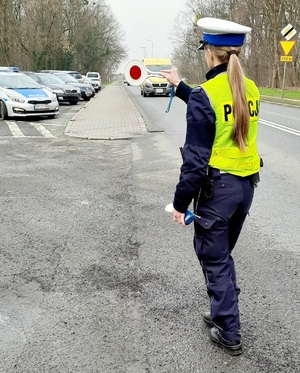 policjantka ruchu drogowego zatrzymująca do kontroli samochód typu bus koloru żółtego, mundurowa trzyma w reku tarcze do zatrzymywania pojazdów oraz urządzenie do sprawdzania stanu trzeźwości