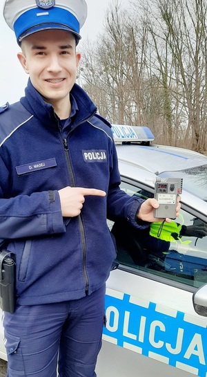 policjant trzymający w ręku urządzenie typu alkosensor, drugą ręką poprzez gest wskazania palcem pokazuje go, w tle radiowóz