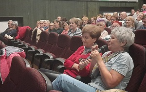 Sala kinowa, widoczna widownia sali kinowej i siedzący na niej seniorzy, kobieta najbliższej obiektywu trzyma w rekach mikrofon