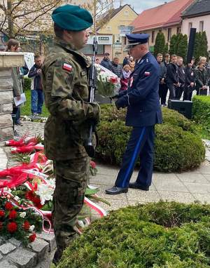 policjant w umundurowaniu galowym składa bukiet kwiatów pod pomnikiem, z lewej strony fotografii widoczny mężczyzna w umundurowaniu żołnierza wojska polskiego