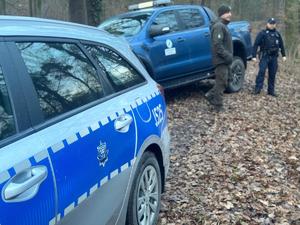 Grafika przedstawia pojazdy policji i straży leśnej. W tle znajduje się las, na terenie którego pełniona jest służba przez funkcjonariuszy obydwu służb.