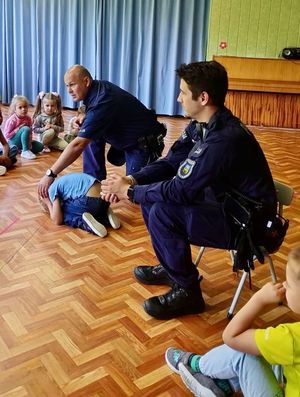 Na fotografii znajdują się Policjanci, którzy pokazują dzieciom pozycję &quot;żółwia&#039;&quot;.