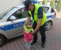 Policjant ubiera dziecku kamizelkę odblaskową, obok stoi radiowóz