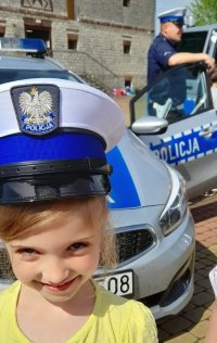 dziewczynka w policyjnej czapce, w tle dwa radiowozy i policjant
