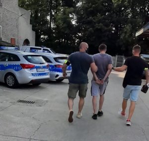 dwaj nieumundurowani policjanci prowadzący zatrzymanego mężczyznę, który ma założone kajdanki na ręce trzymane do tyłu, w tle dwa radiowozy oznakowane i budynek Komendy
